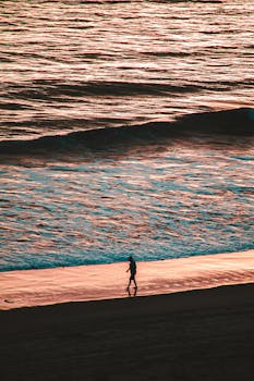 日落时分，一个孤独的人沿着沙滩散步，身影在海浪中留下轮廓