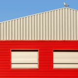 A modern industrial building with a red facade and a seagull perched on the roof under clear blue skies.