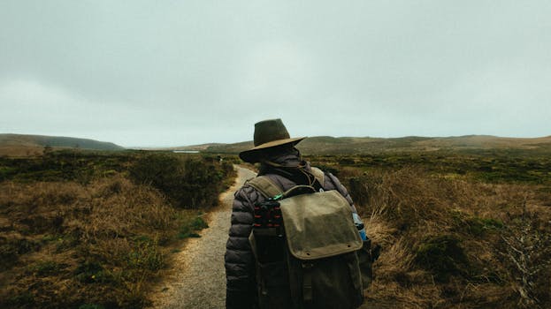 一名徒步旅行者头戴帽子、背着背包，沿着薄雾缭绕的乡村小路行走，探索大自然