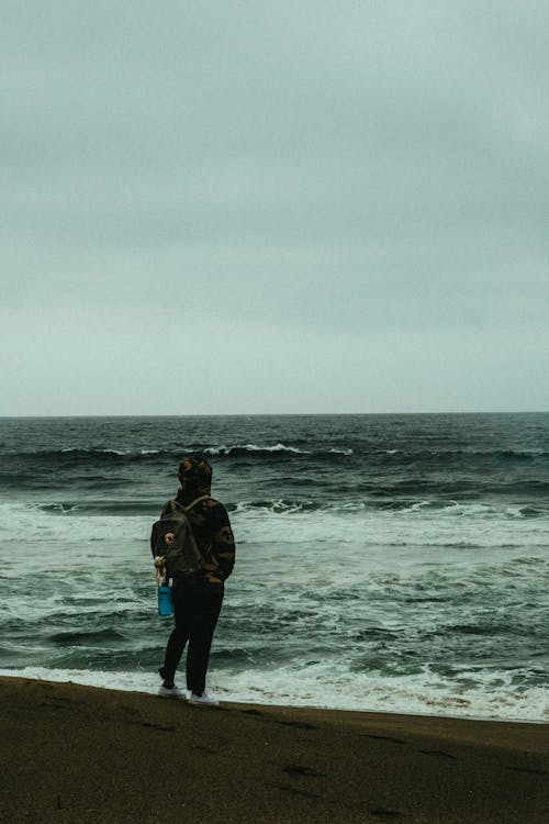 Photo of Person Carrying Backpack Standing Alone on the Beach Overlooking the Horizon