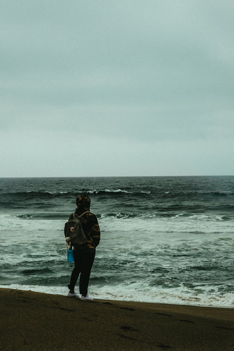 Photo Of Person Carrying Backpack Standing Alone On The Beach Overlooking The Horizon