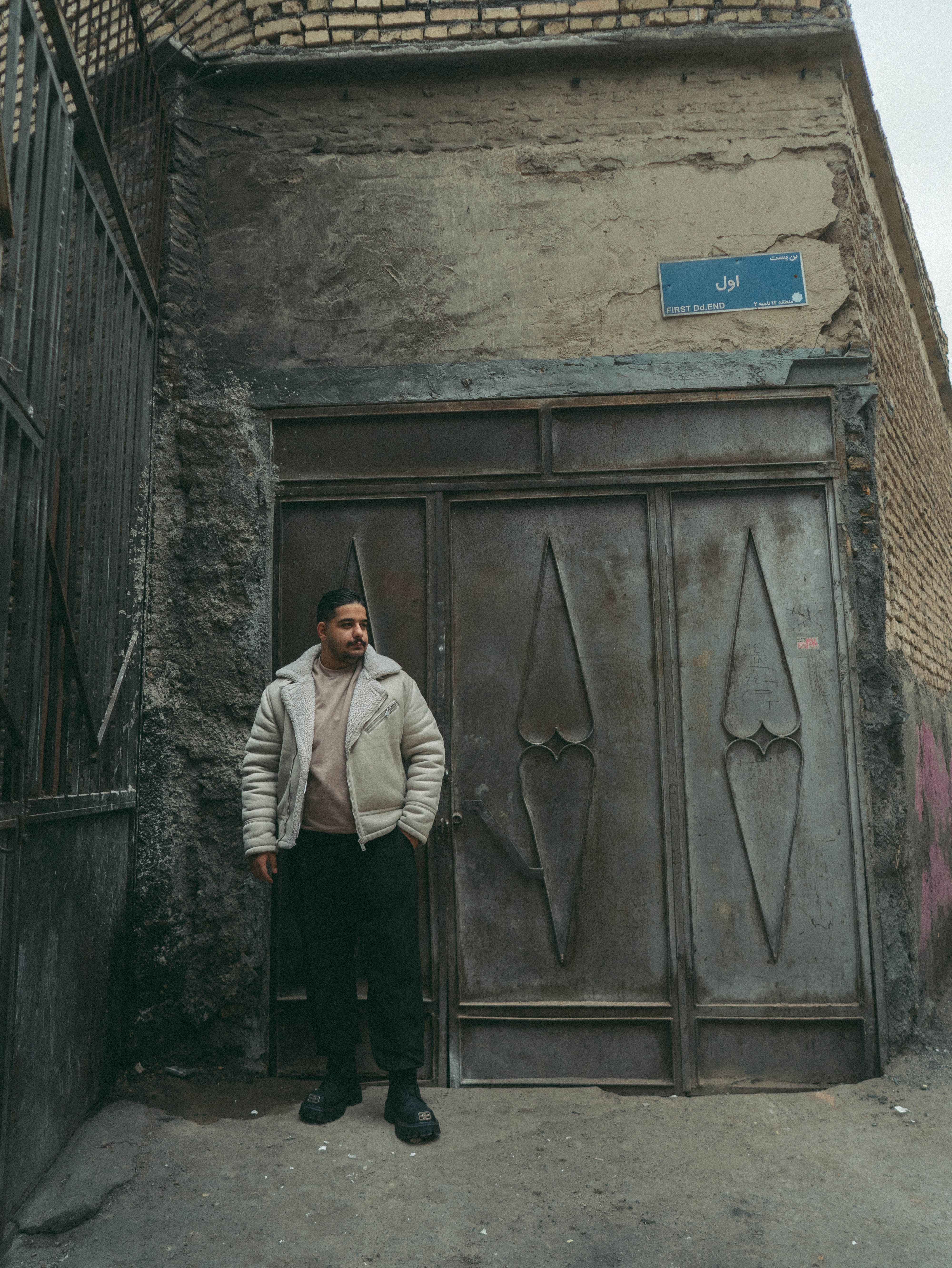 A man stands quietly by a rustic metal door in a narrow urban alleyway.