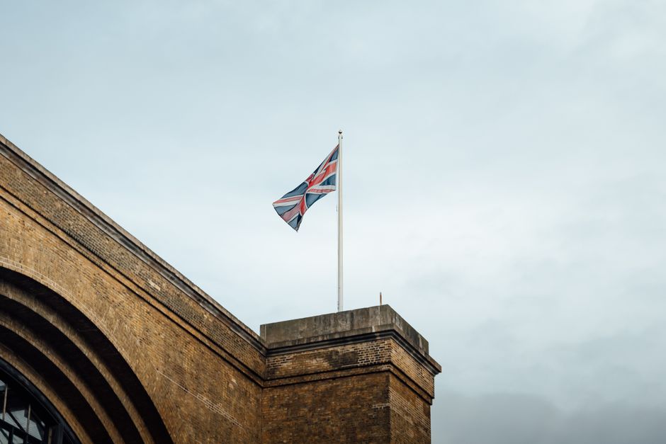 Union Jack flag waving atop a historic brick building in London, UK, against a cloudy sky.