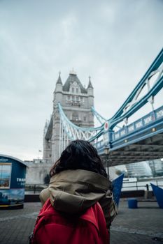 A traveler with a red backpack gazes at the iconic Tower Bridge in London on a cloudy day.