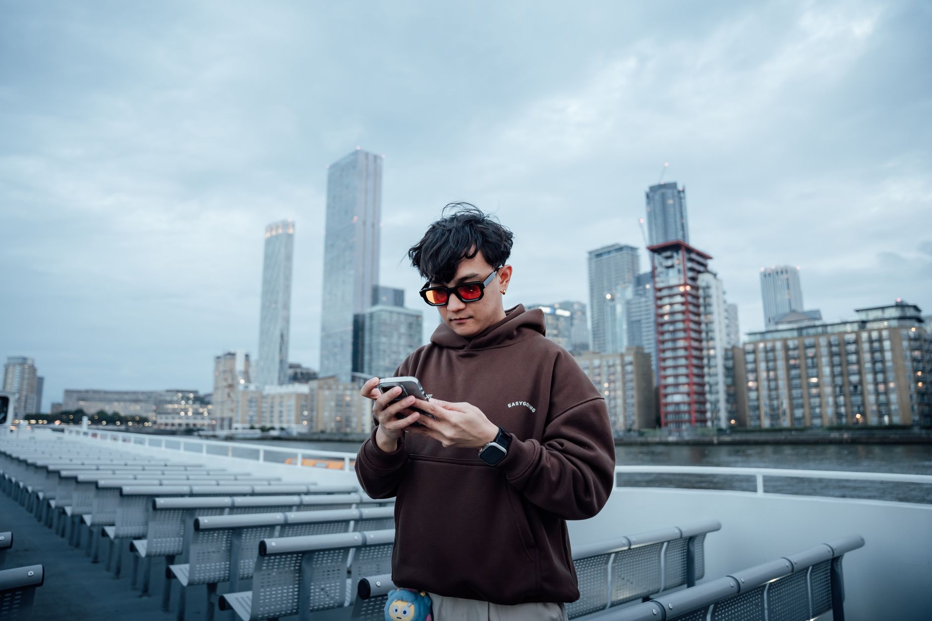 Casual young man in sunglasses using smartphone on a riverboat in London with city skyline background.