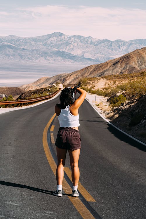 Back View Photo of Woman in White Tank Top and Brown Shorts Standing in the Middle of an Empty Road Taking a Photo 