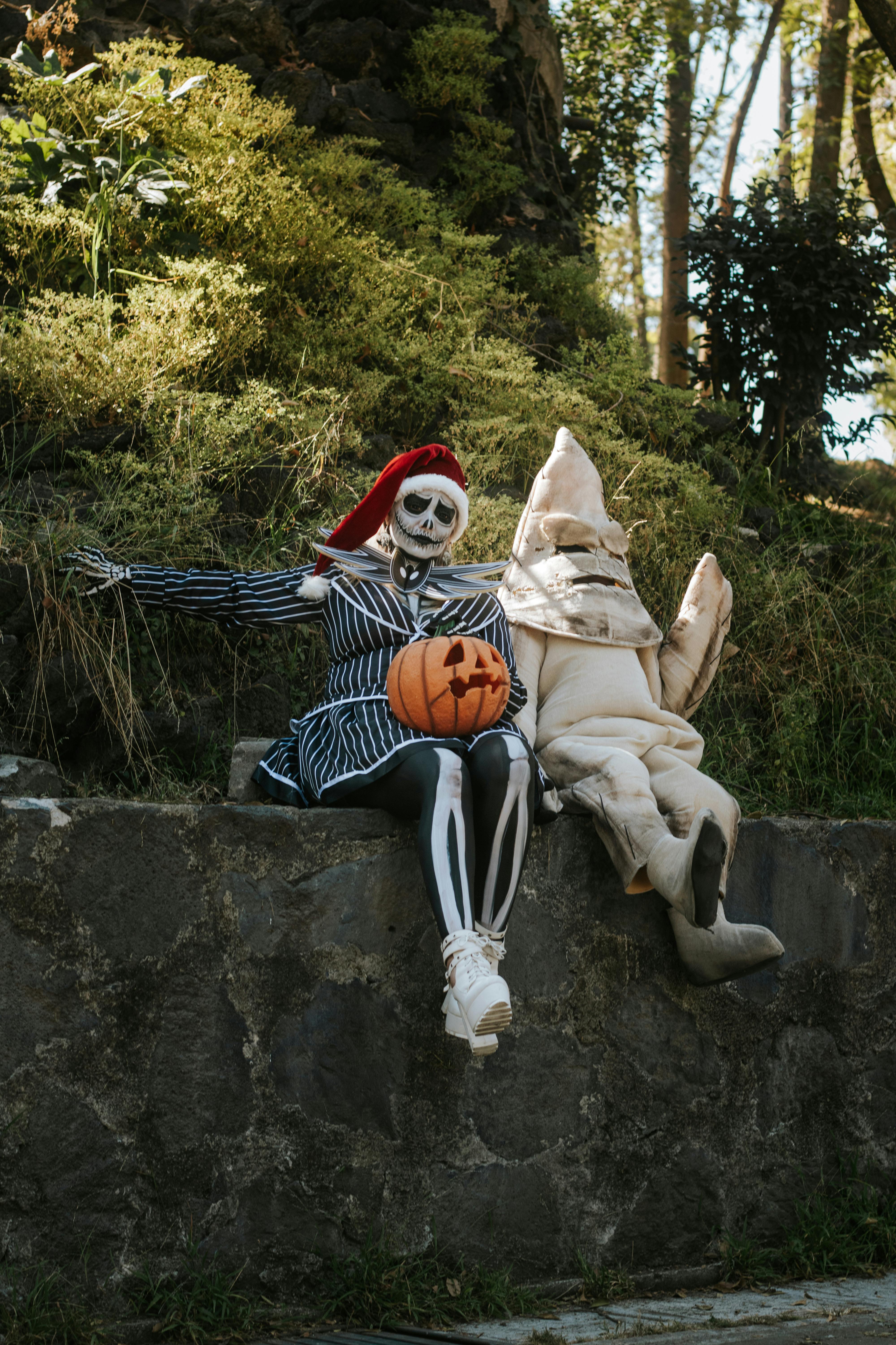Two people in Halloween costumes sitting outdoors with a pumpkin.