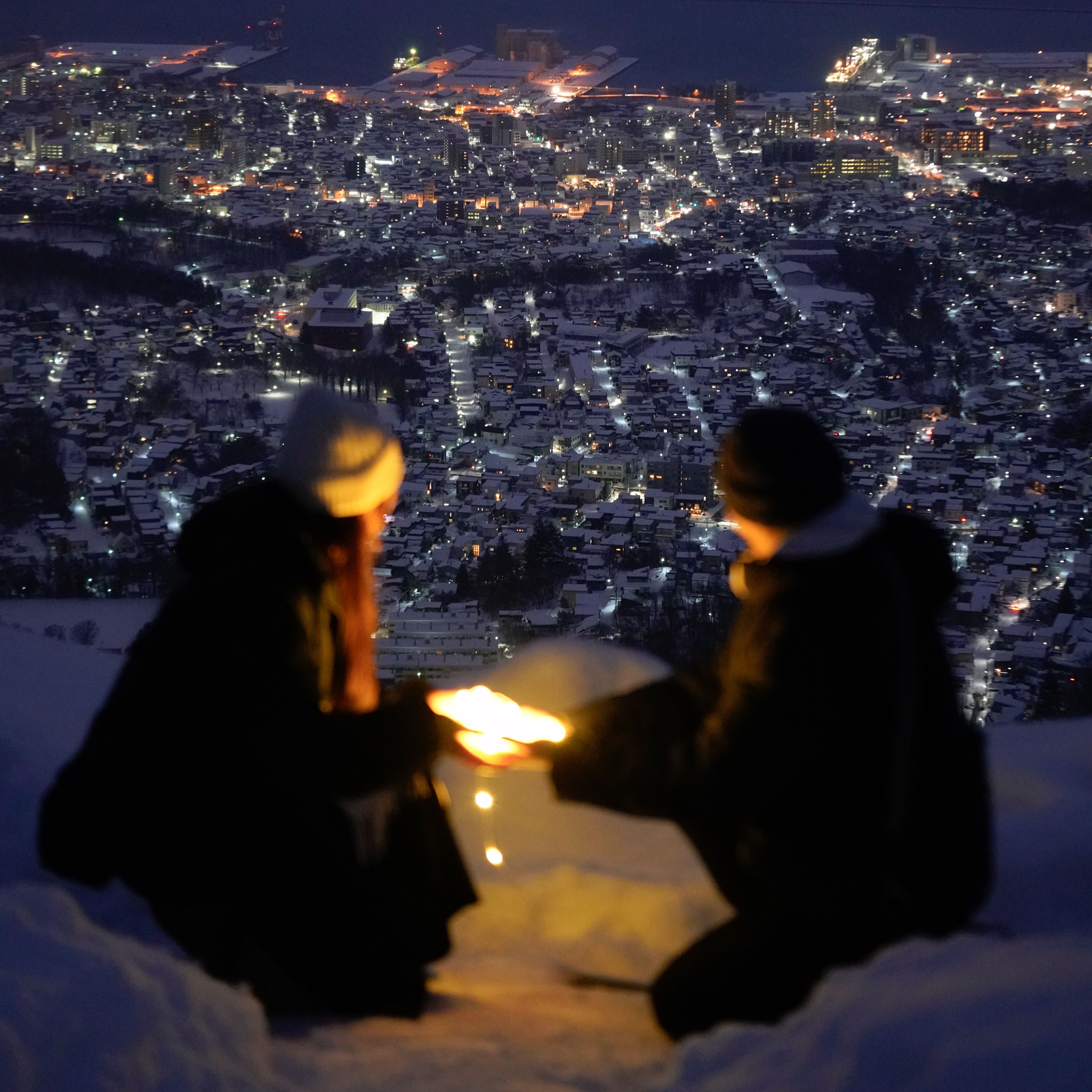 Two adults share a warm moment with candles above a snow-covered city at night.