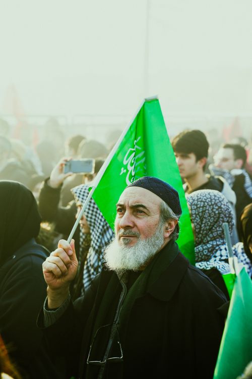 Free An elderly man with a beard holding a green flag at a public gathering, surrounded by people. Stock Photo