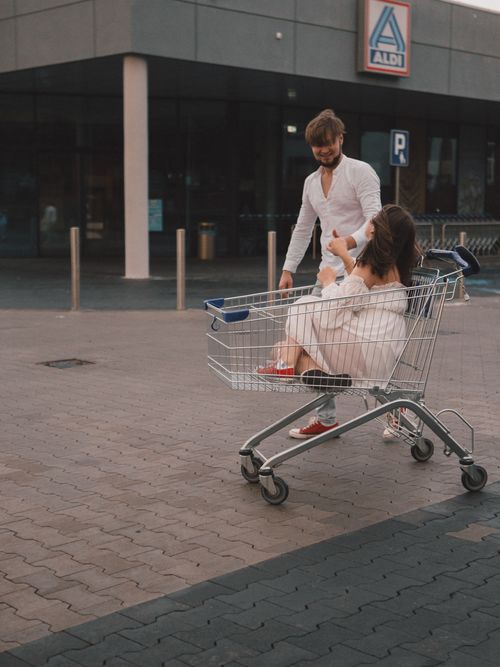 Free Couple enjoying a playful moment with a shopping cart outside a supermarket. Stock Photo