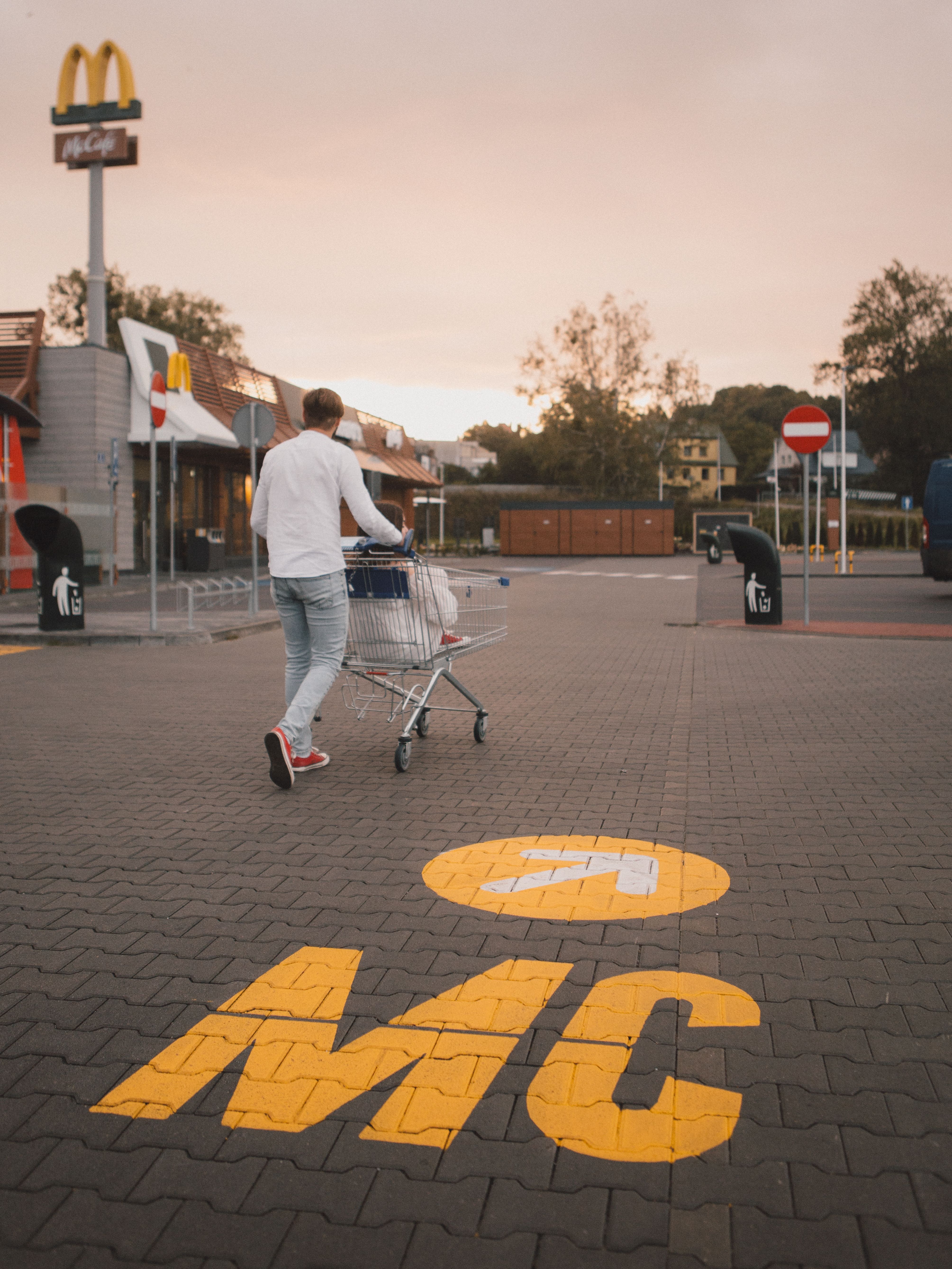 A person walks with a shopping cart in an outdoor fast food restaurant parking lot at sunset.