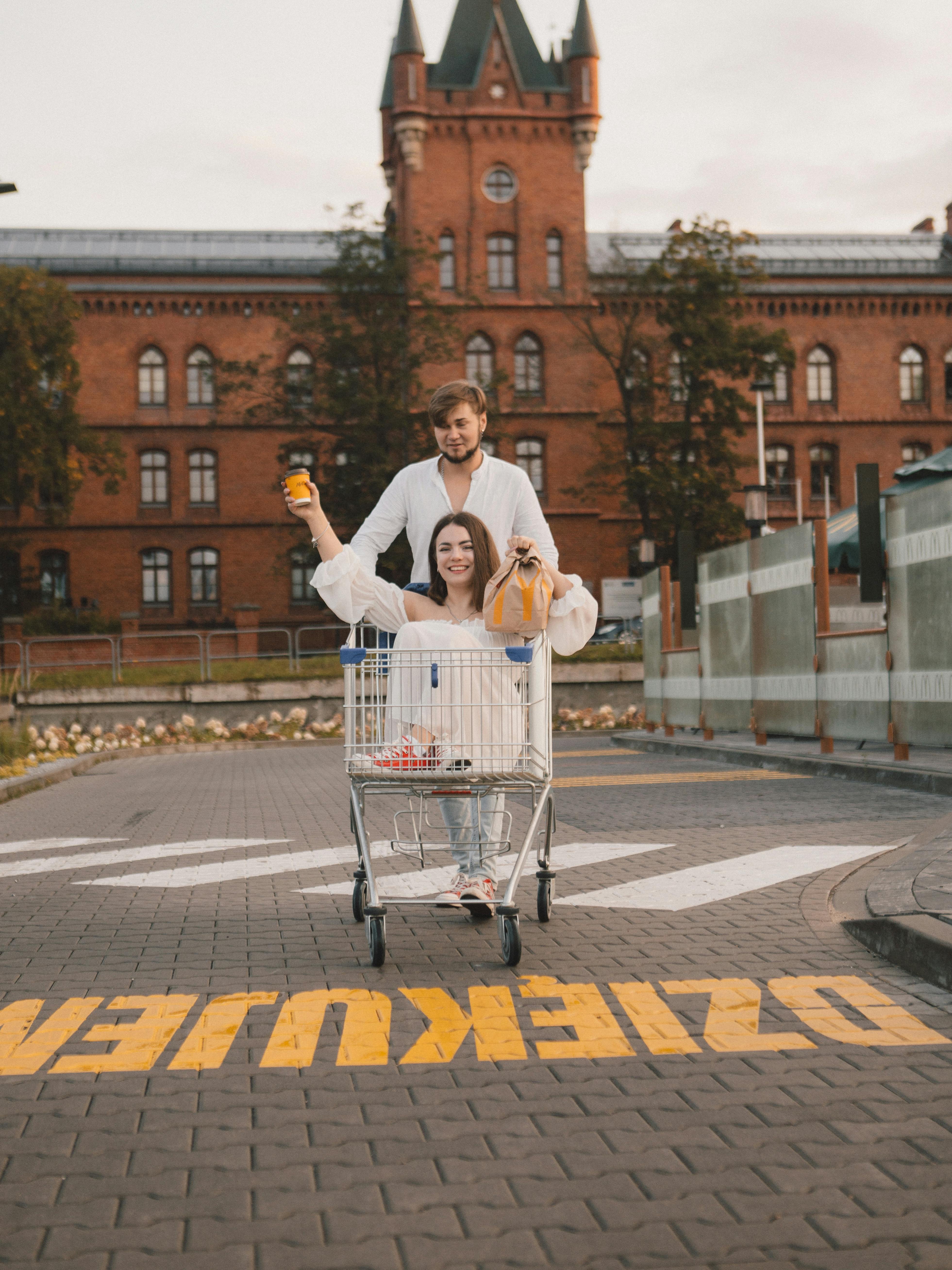 Happy couple enjoys a playful moment in a shopping cart in front of a historic building.