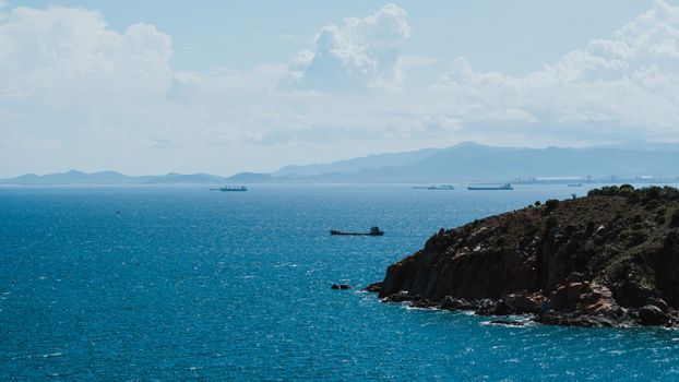 A tranquil ocean scene with ships on the horizon and a rugged, rocky coastline under a bright sky.