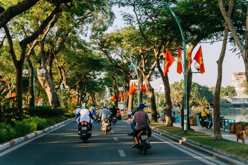 Free Motorbikes ride along a tree-lined street with flags in Hanoi, Vietnam during sunset. Stock Photo
