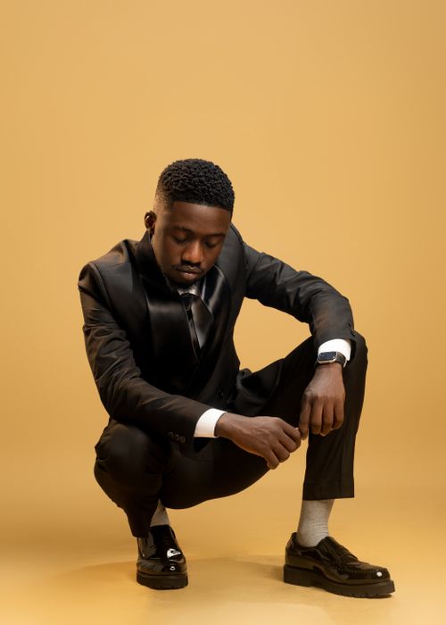 Free Elegant young man in a black suit crouching against a warm, neutral backdrop. Stock Photo