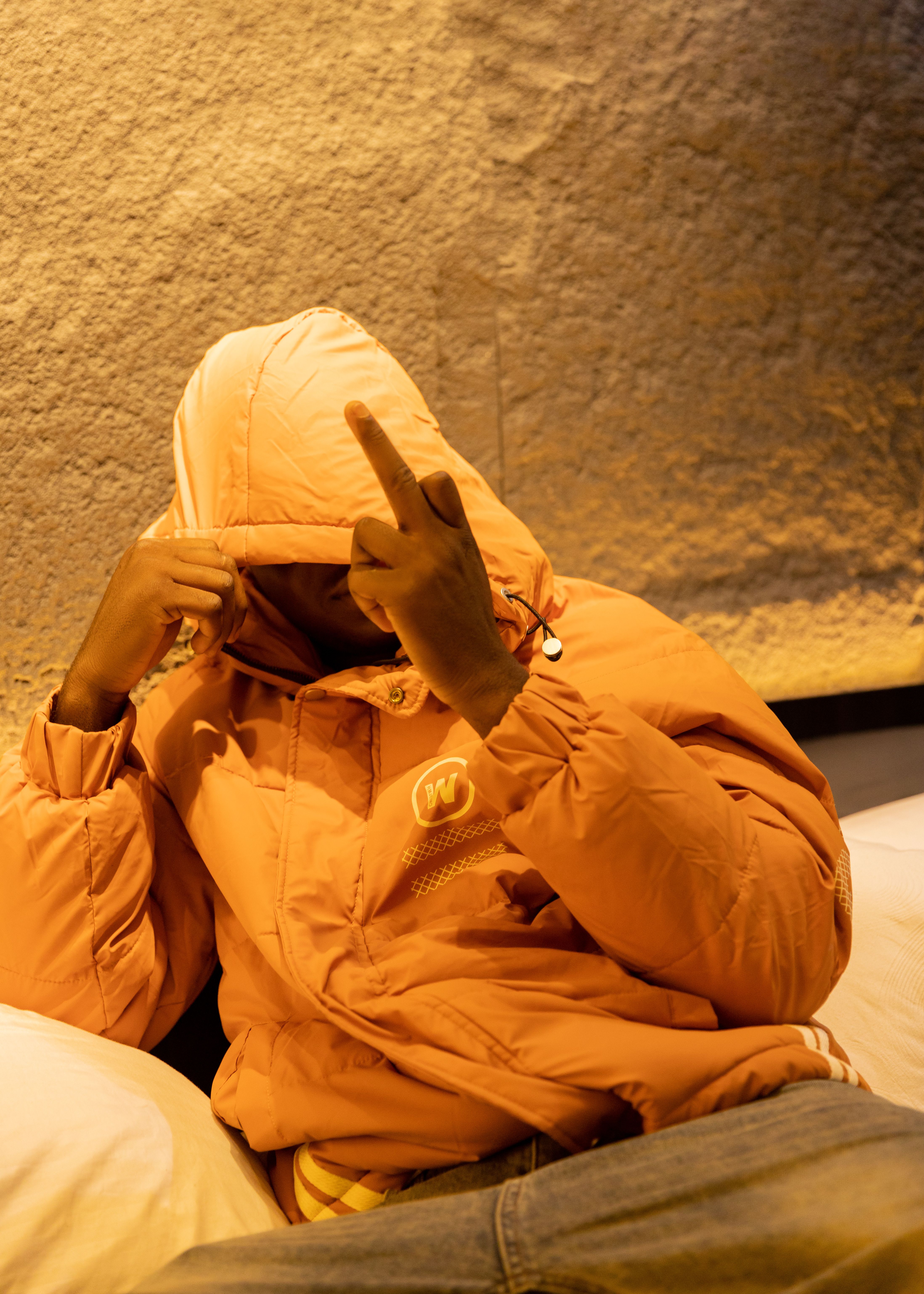 Person in orange jacket partially covering face with hood, sitting relaxed indoors.