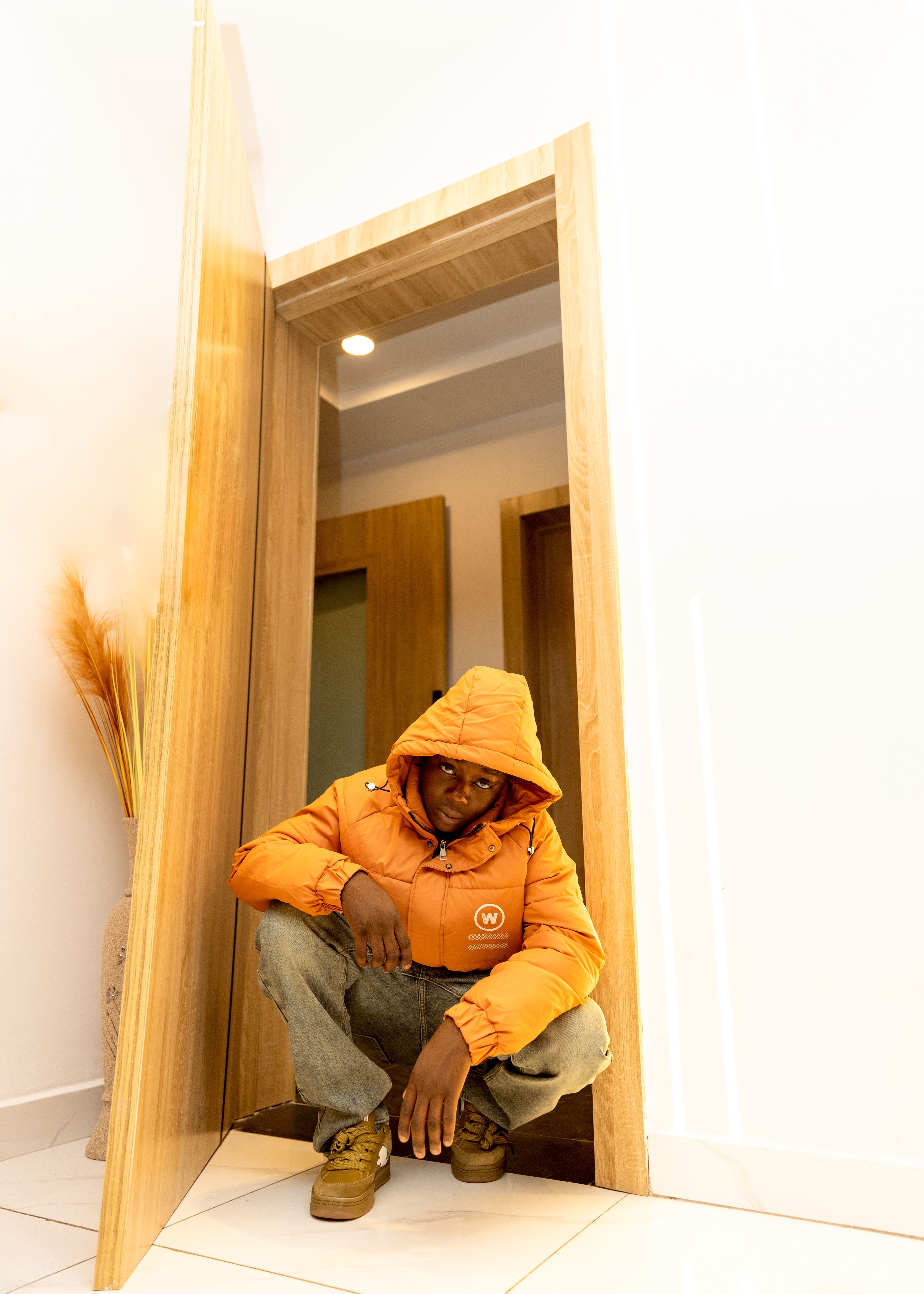 Man squatting casually in doorway wearing an orange jacket in a modern indoor setting.