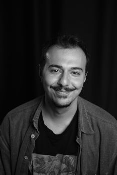 Monochrome studio portrait of a stylish bearded man in casual attire with a confident smile.