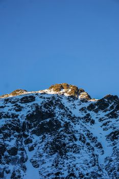 A majestic snow-covered mountain peak illuminated by the sun against a clear blue sky.