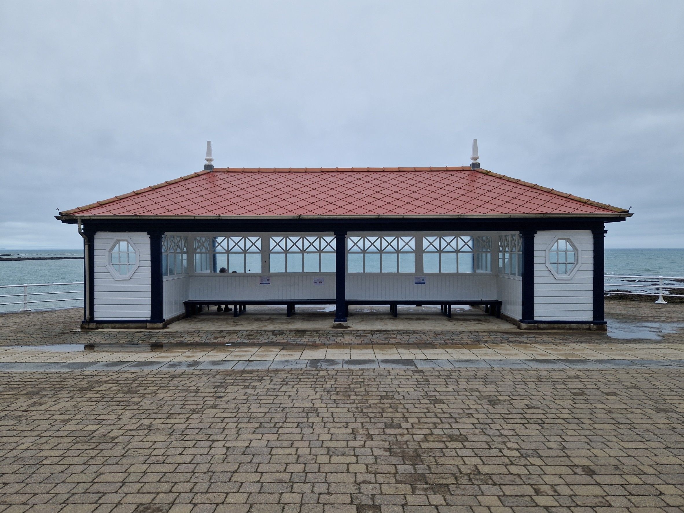 Seafront shelter in Aberystwyth, United Kingdom, with a red tiled roof, distant people, grey skies and the sea beyond