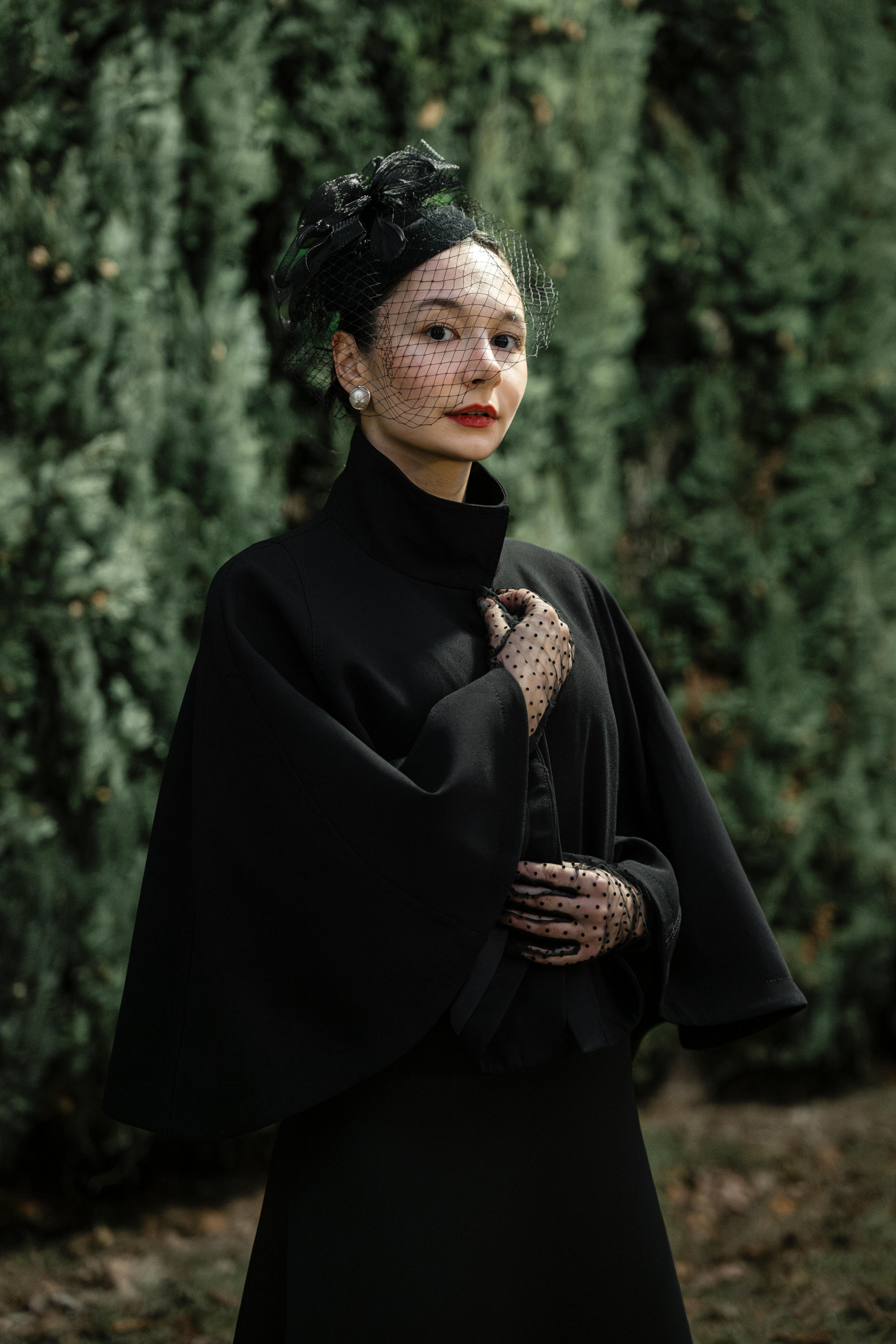 Free Portrait of an elegant woman in a vintage black outfit with netted hat against a lush green background. Stock Photo