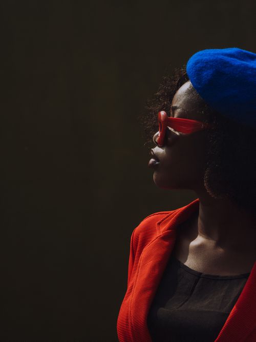 Free A fashionable woman in a red jacket and blue hat, wearing sunglasses, against a dark backdrop. Stock Photo
