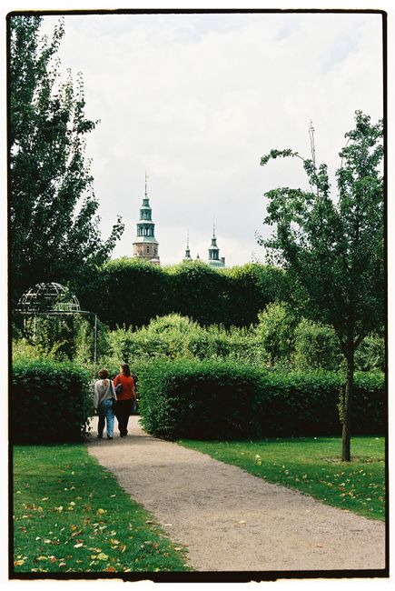 Scenic walkway framed by lush greenery leading to distant spires under a cloudy sky.