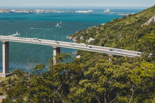 Free Stunning aerial view of a bridge spanning clear blue waters with lush green hills and a vibrant coastline backdrop. Stock Photo