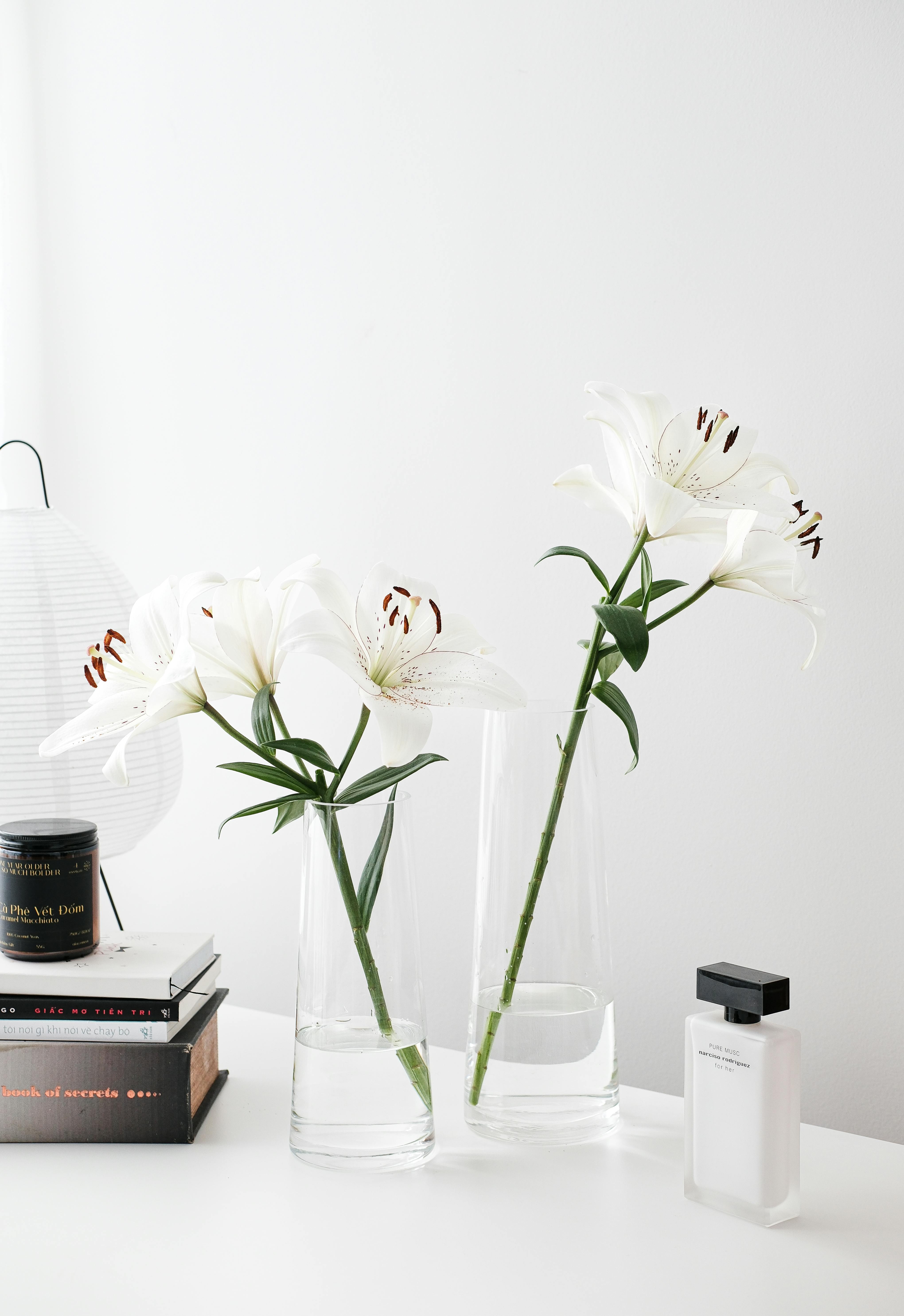 Stylish white lilies in glass vases enhance a modern minimalist desk setup with books and perfume.