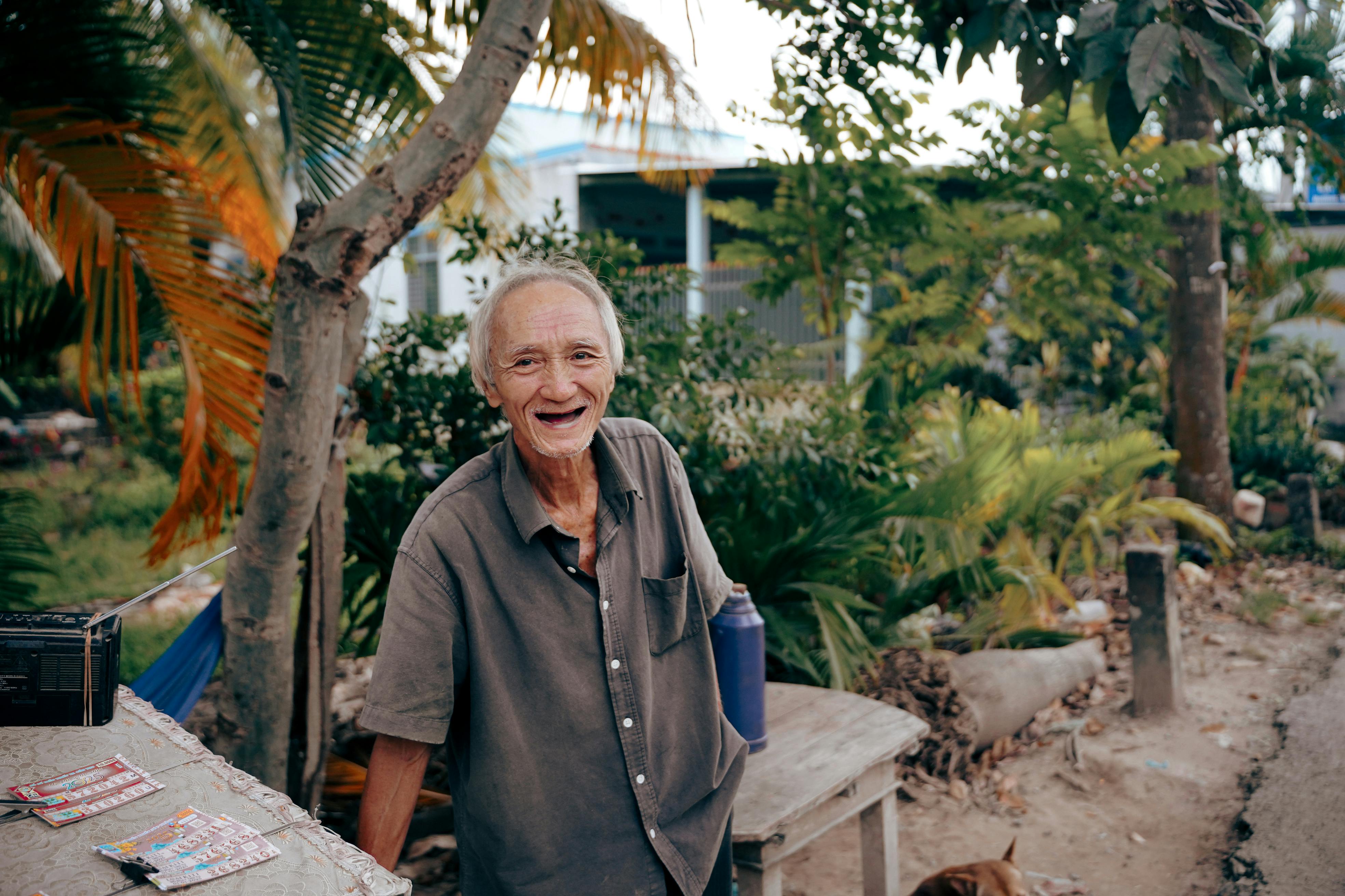 A joyful elderly man shares a smile outdoors in a garden in Bến Tre, Vietnam, surrounded by lush greenery.
