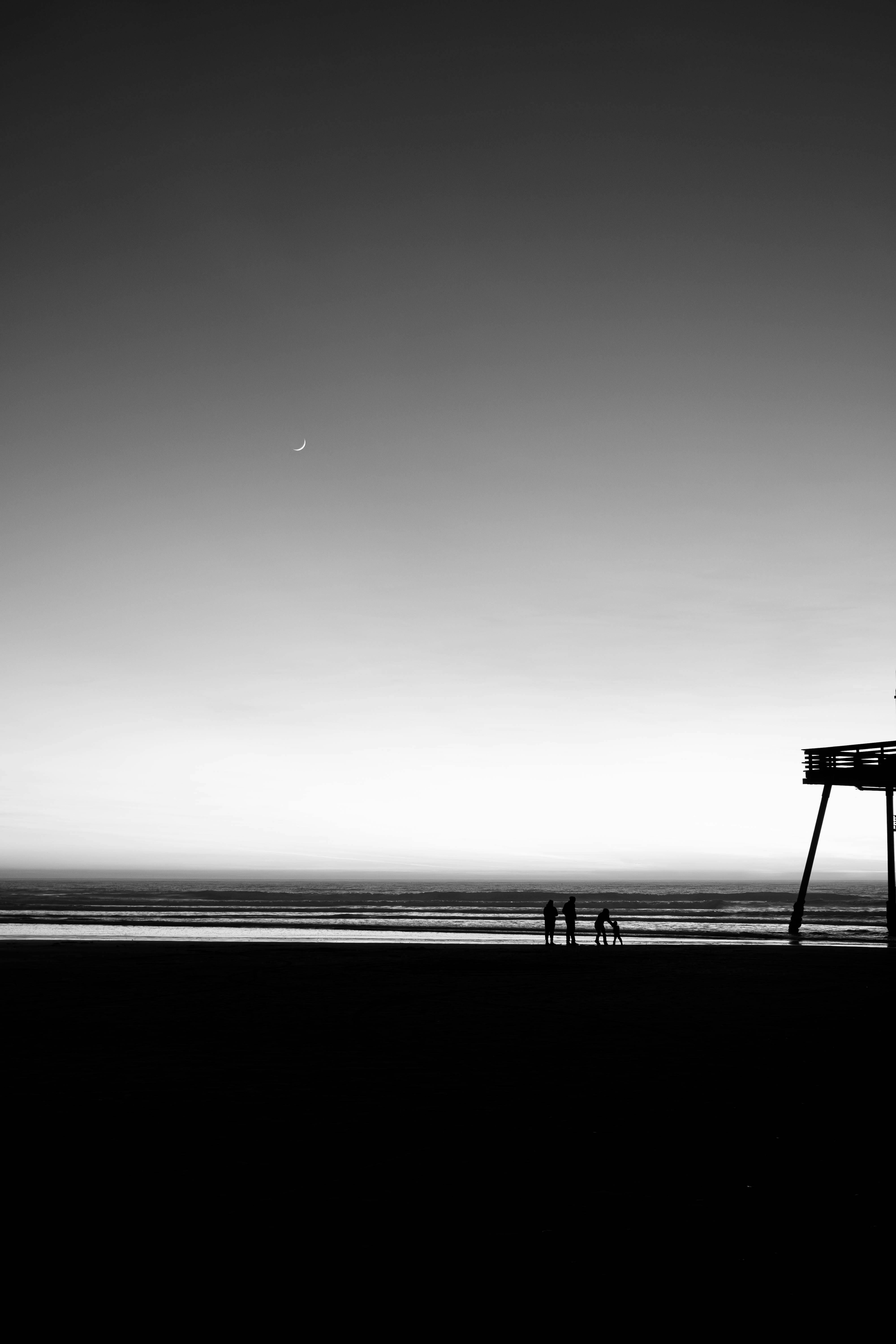 A serene black and white image of a beach with a crescent moon and pier at dusk.