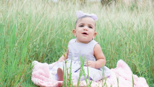 Free Charming baby girl in a dress with a bow headband sitting on a blanket amid tall grass. Stock Photo