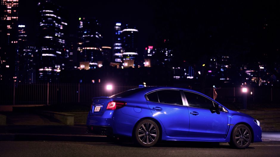 A blue Subaru WRX parked with a vibrant city skyline backdrop, captured during night time.