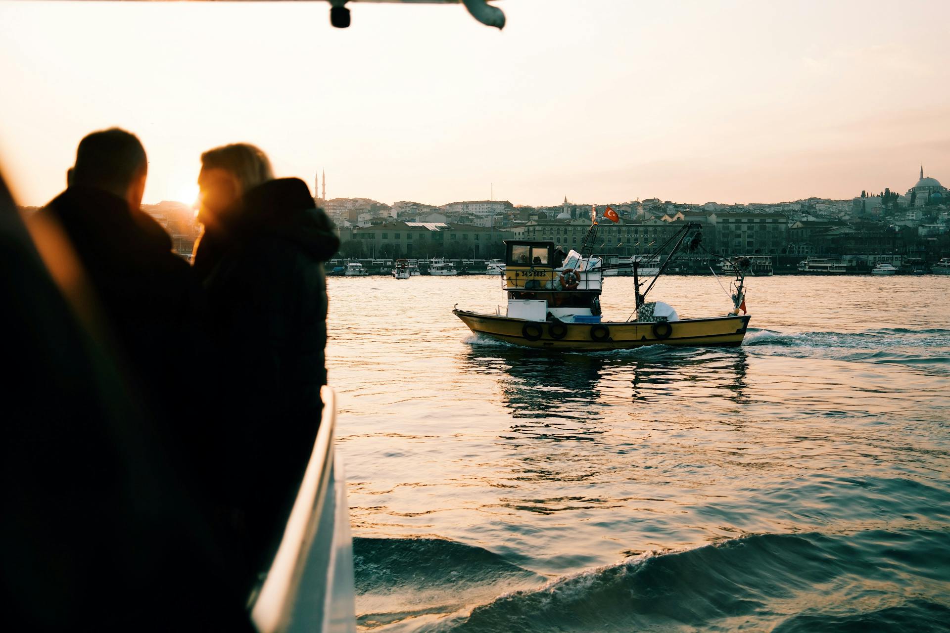 Silhouetted figures on a ferry with a scenic Istanbul sunset and fishing boat in view.