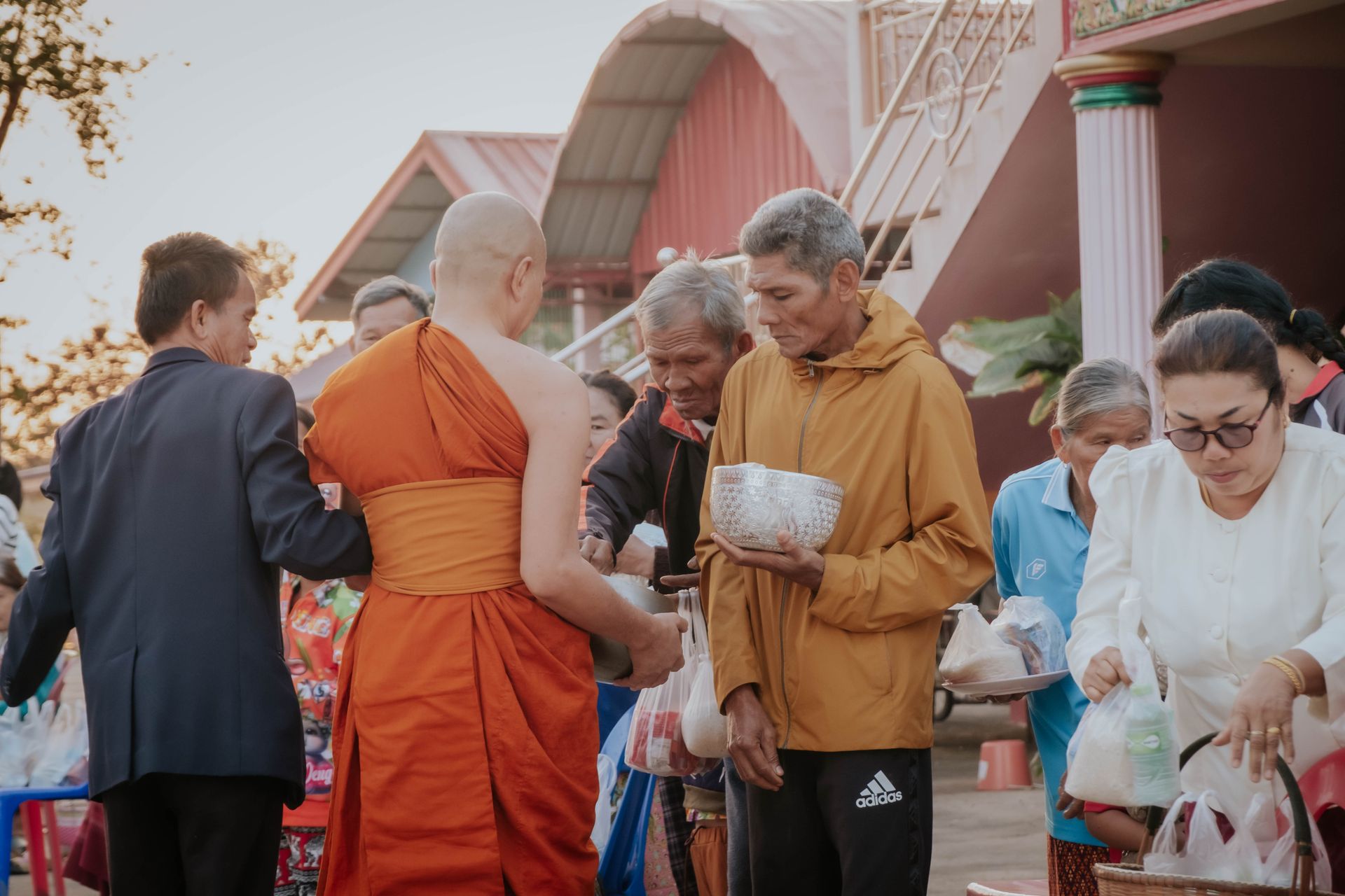 A group of people giving alms to Buddhist monks during a religious ceremony outdoors, in traditional setting.