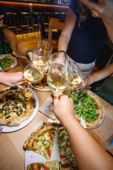 Friends clinking glasses over gourmet pizzas in a lively restaurant setting.