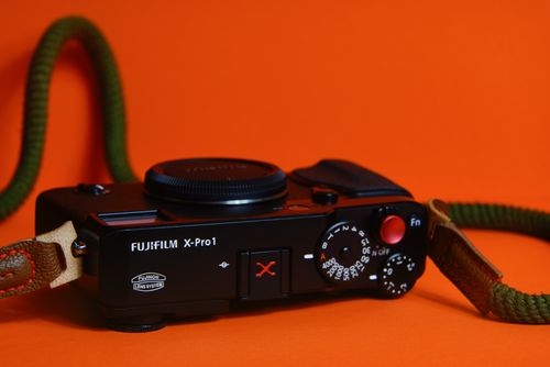 Free Close-up of a retro Fujifilm X-Pro1 camera with a strap against a vibrant orange background. Stock Photo