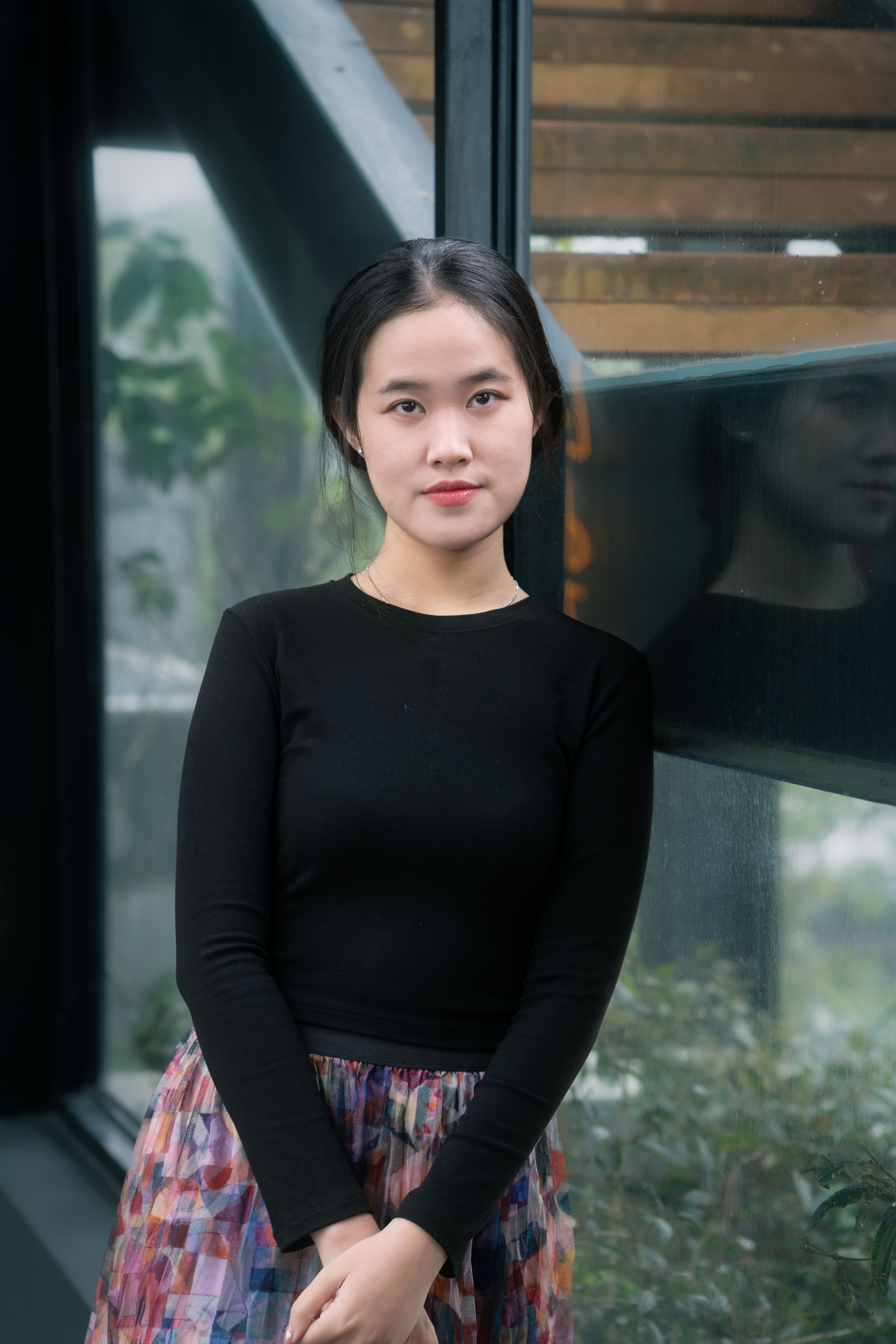 Portrait of a young woman in a black top, standing by a modern indoor window with greenery outside.