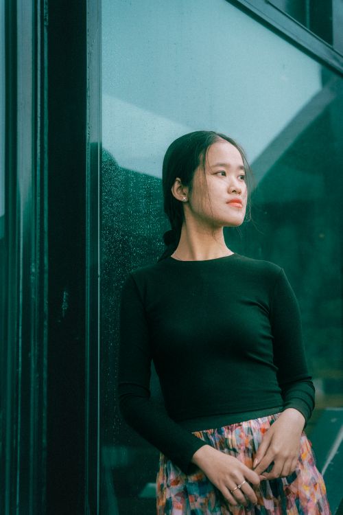 Free A young woman in a black top stands pensively against a glass wall, looking away. Stock Photo