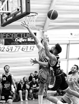 Black and white photo captures a thrilling moment in an indoor basketball game with players reaching for the hoop.