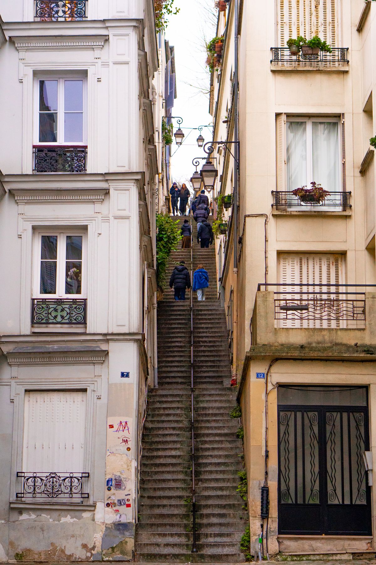 Rue en escalier de Montmartre bordée d’immeubles à volets, longue volée de marches et réverbères, quelques passants de dos gravissant les marches