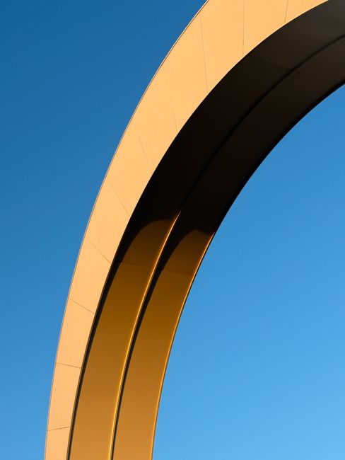 A sleek gold arch structure stands against a bright blue sky, showcasing modern design elements.