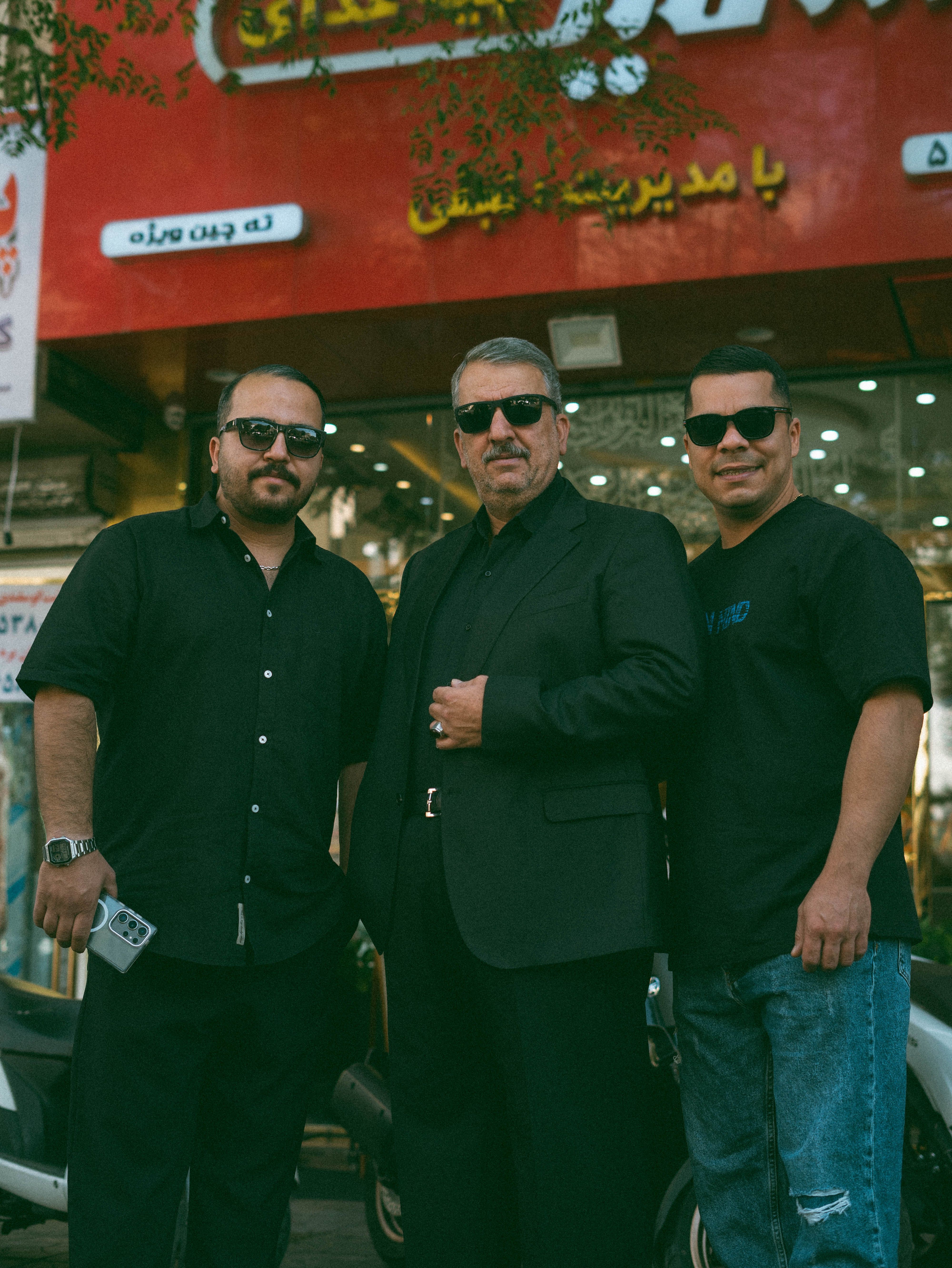 Three men in black sunglasses stand confidently outdoors. Urban background with signage.