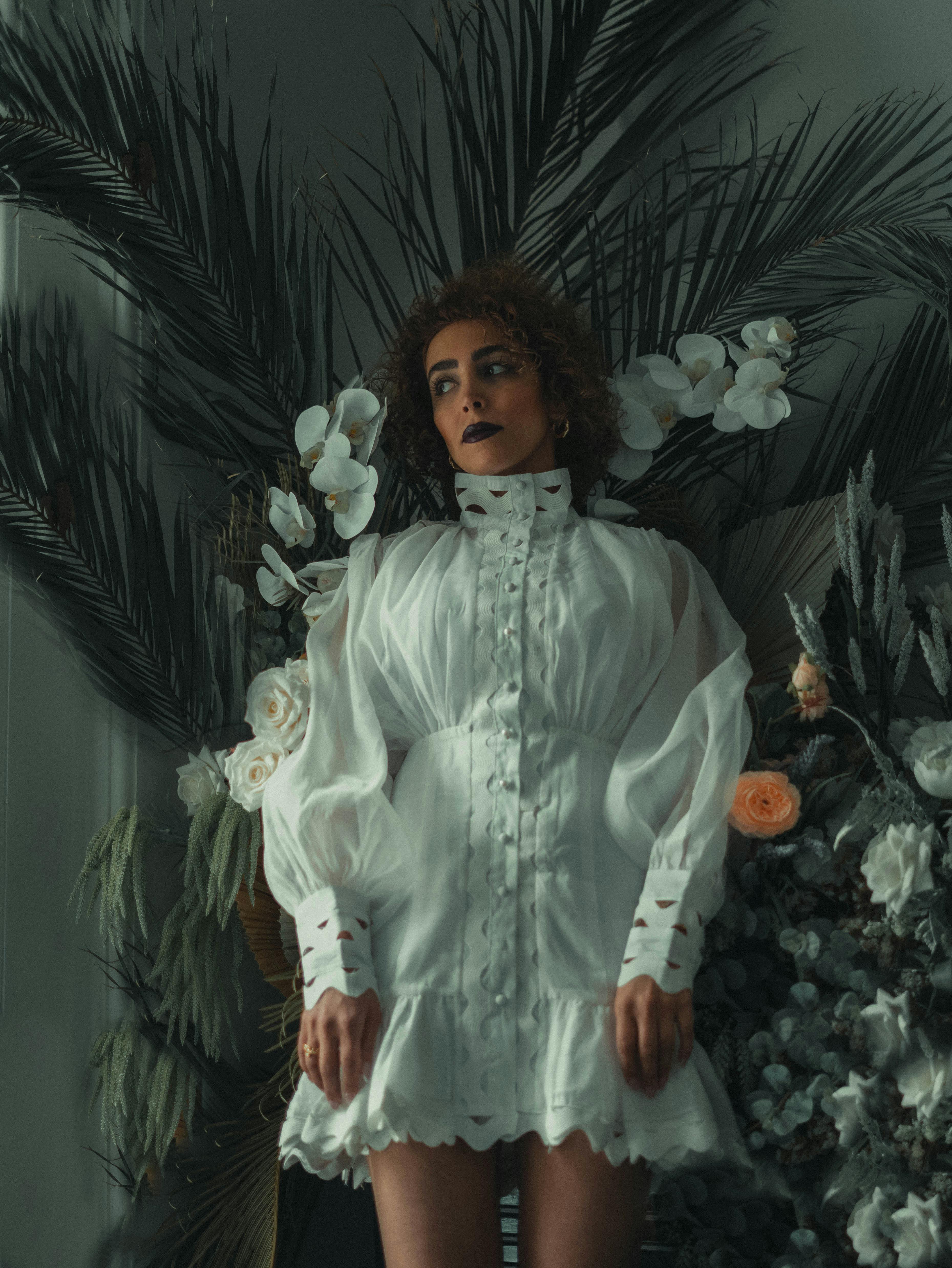 A woman in a white dress stands gracefully among lush foliage and flowers.