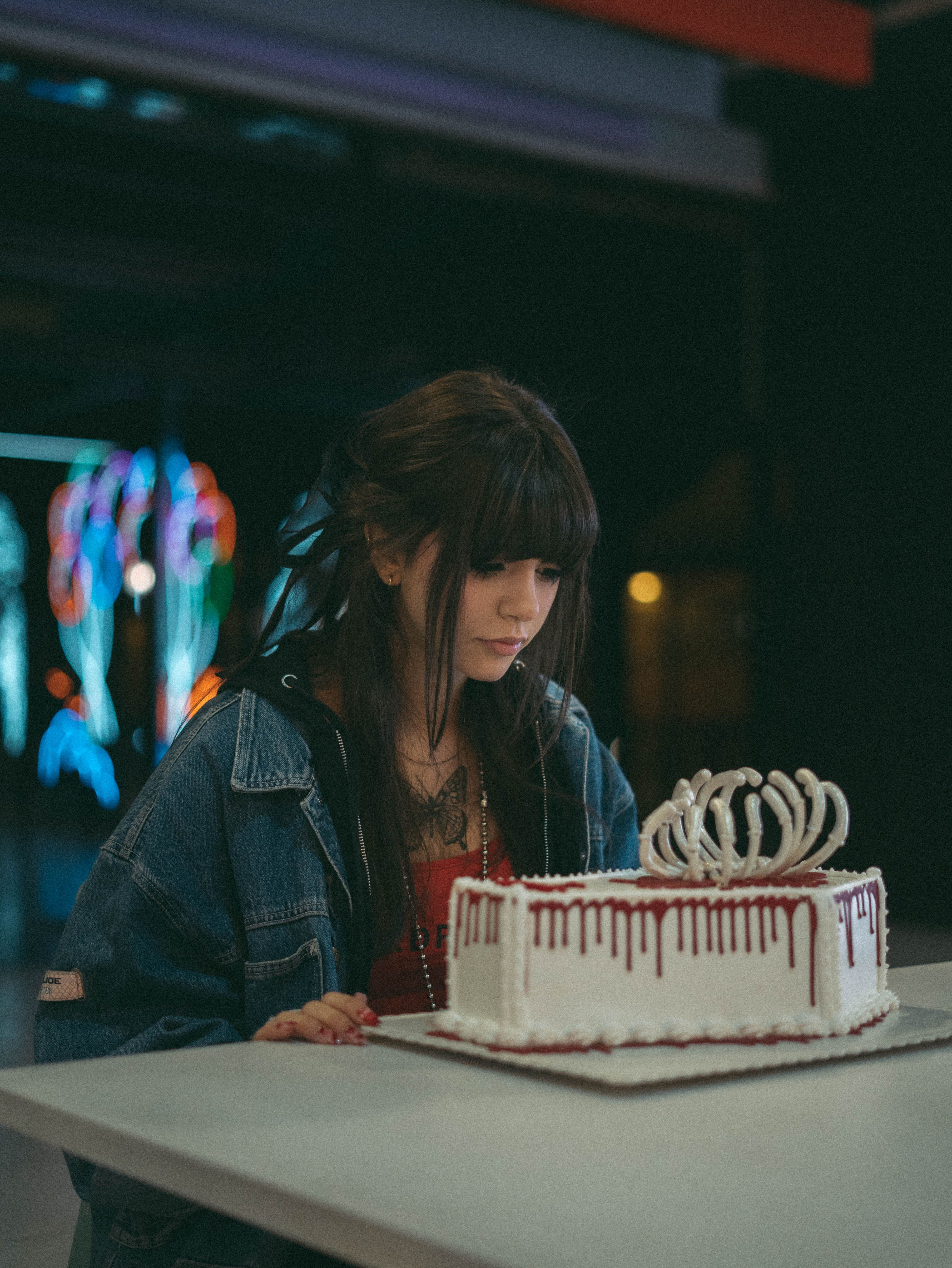 Woman with dark hair sits alone at table with artistic cake in dimly lit setting.