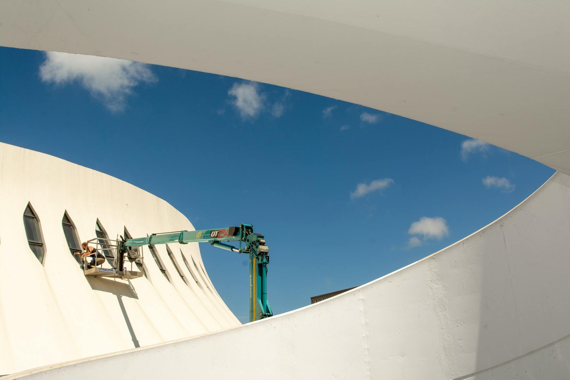 A crane conducting maintenance on a unique modern building with sweeping lines under a clear blue sky.