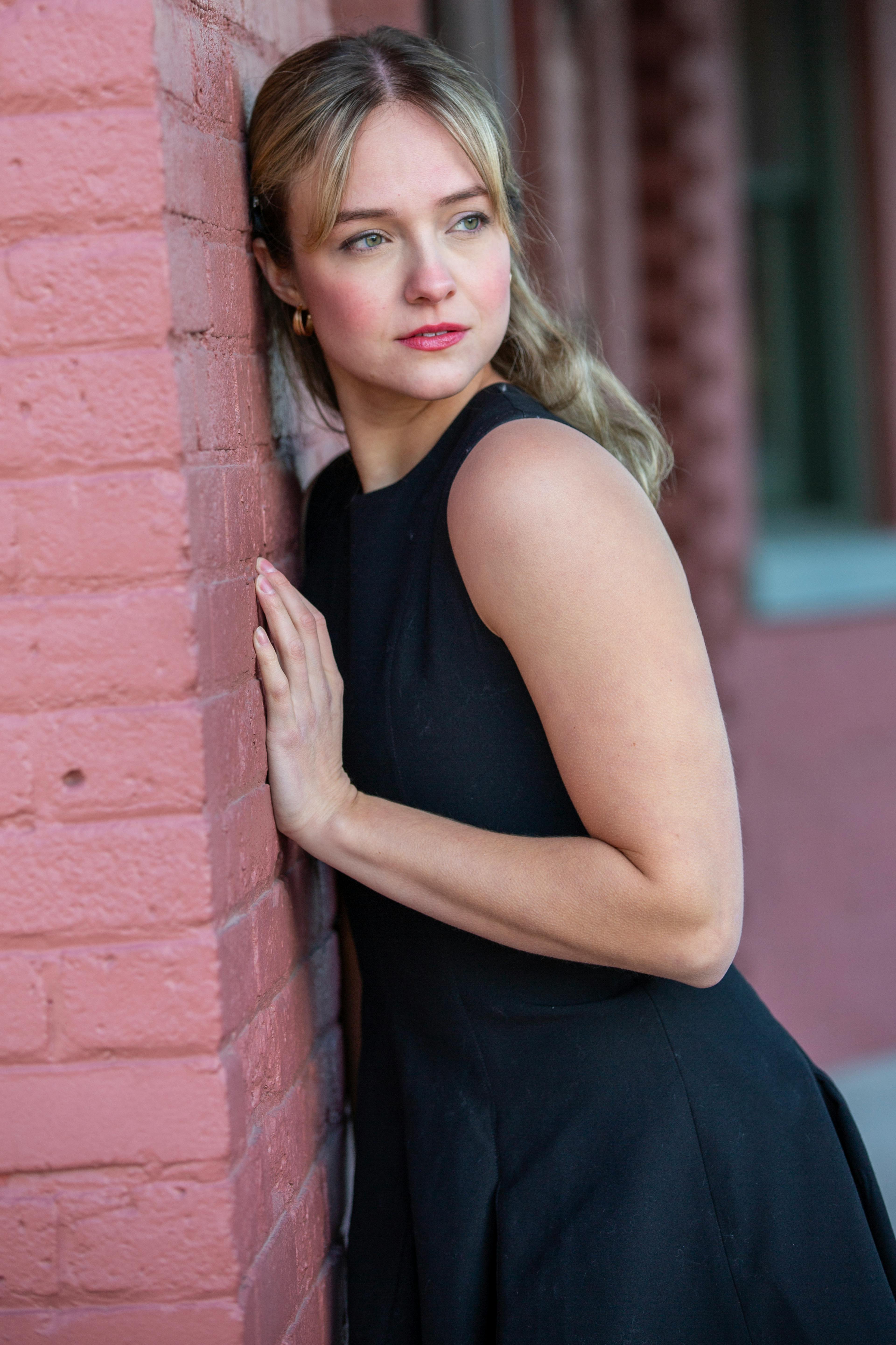 Stylish woman in a black dress poses against a red brick wall outdoors.