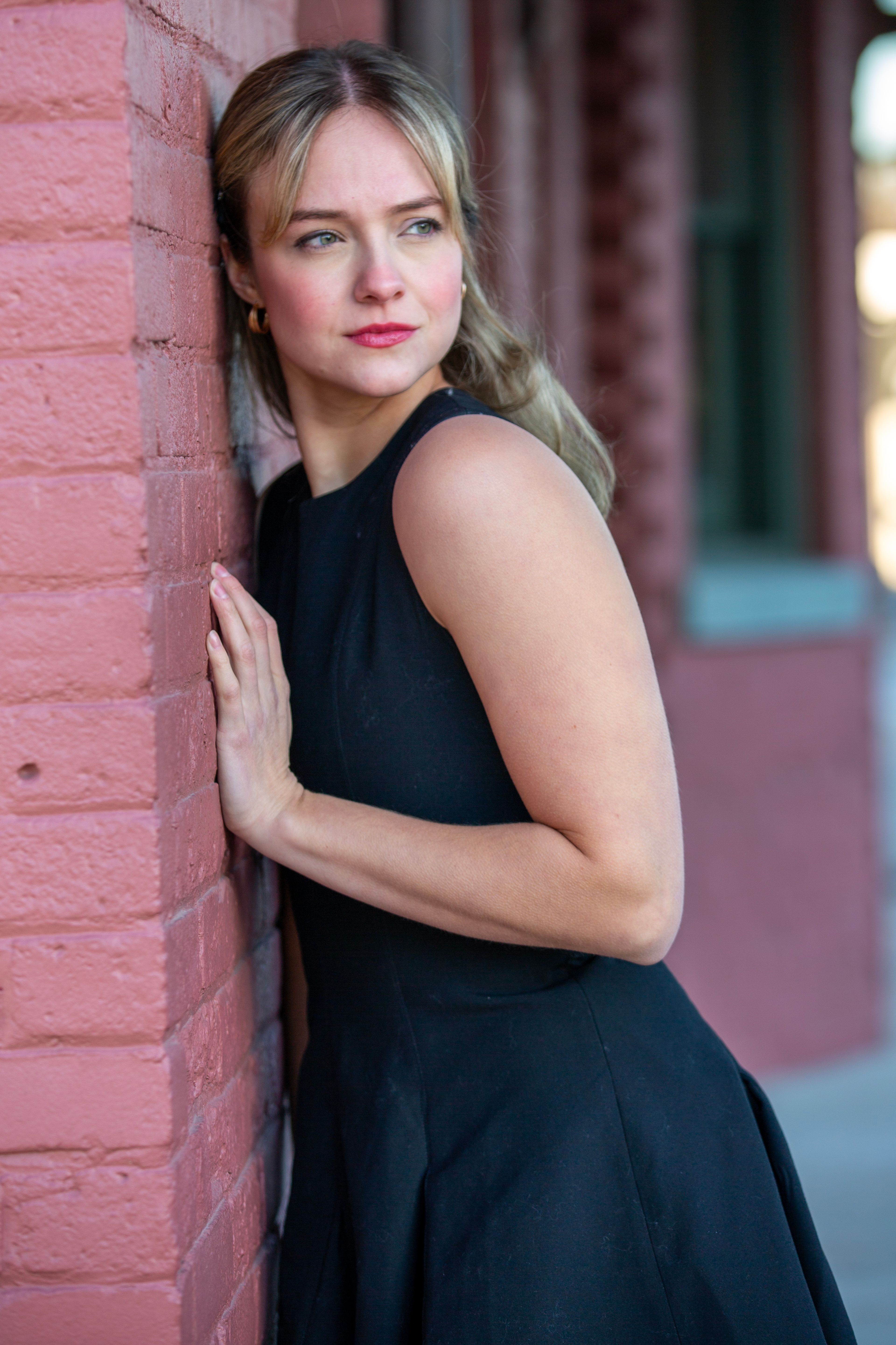 A woman in a black dress leans against a red brick wall, exuding elegance and contemplation.