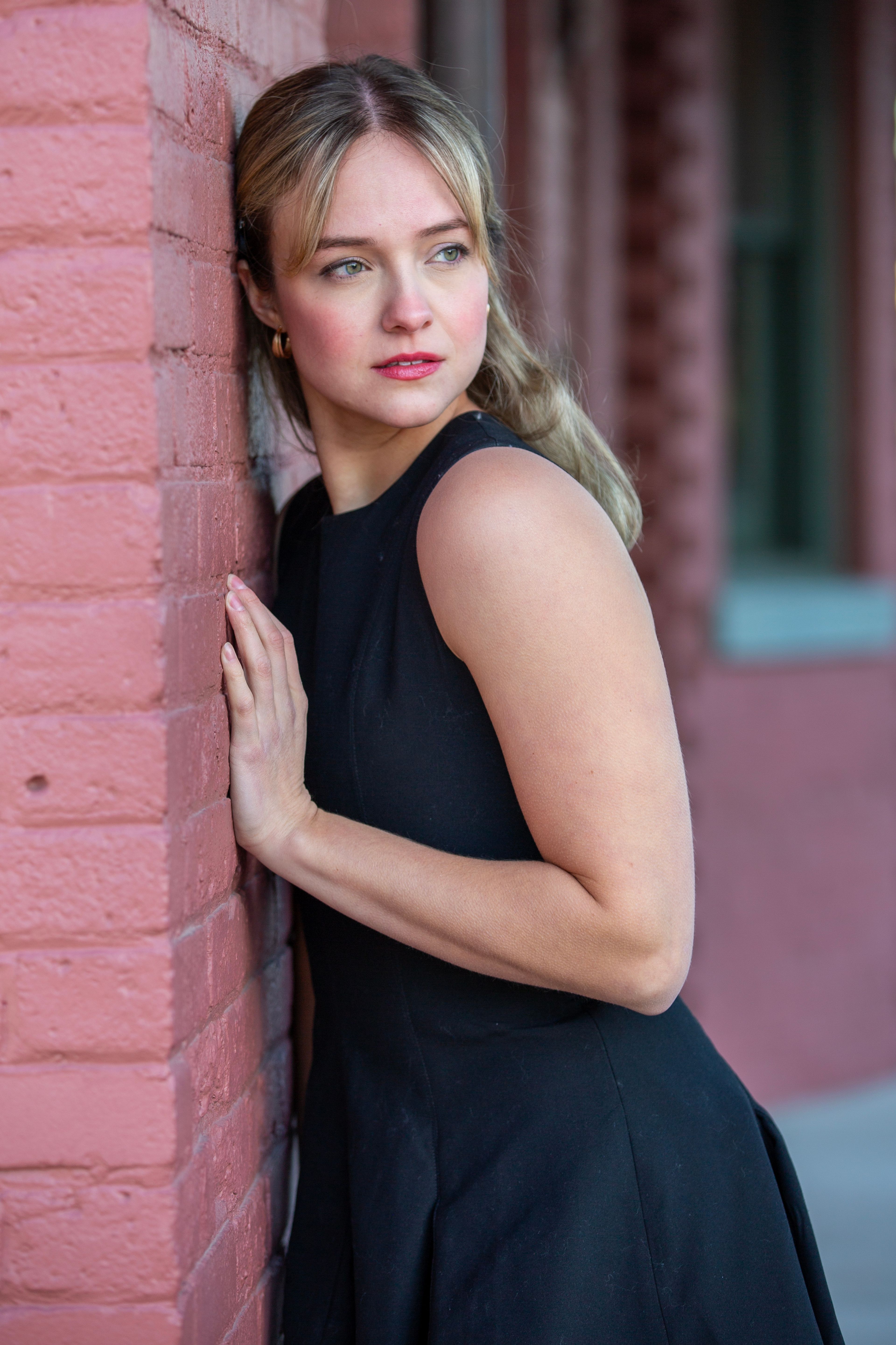 Stylish woman in black dress leans on brick wall with thoughtful expression. Urban fashion portrait.