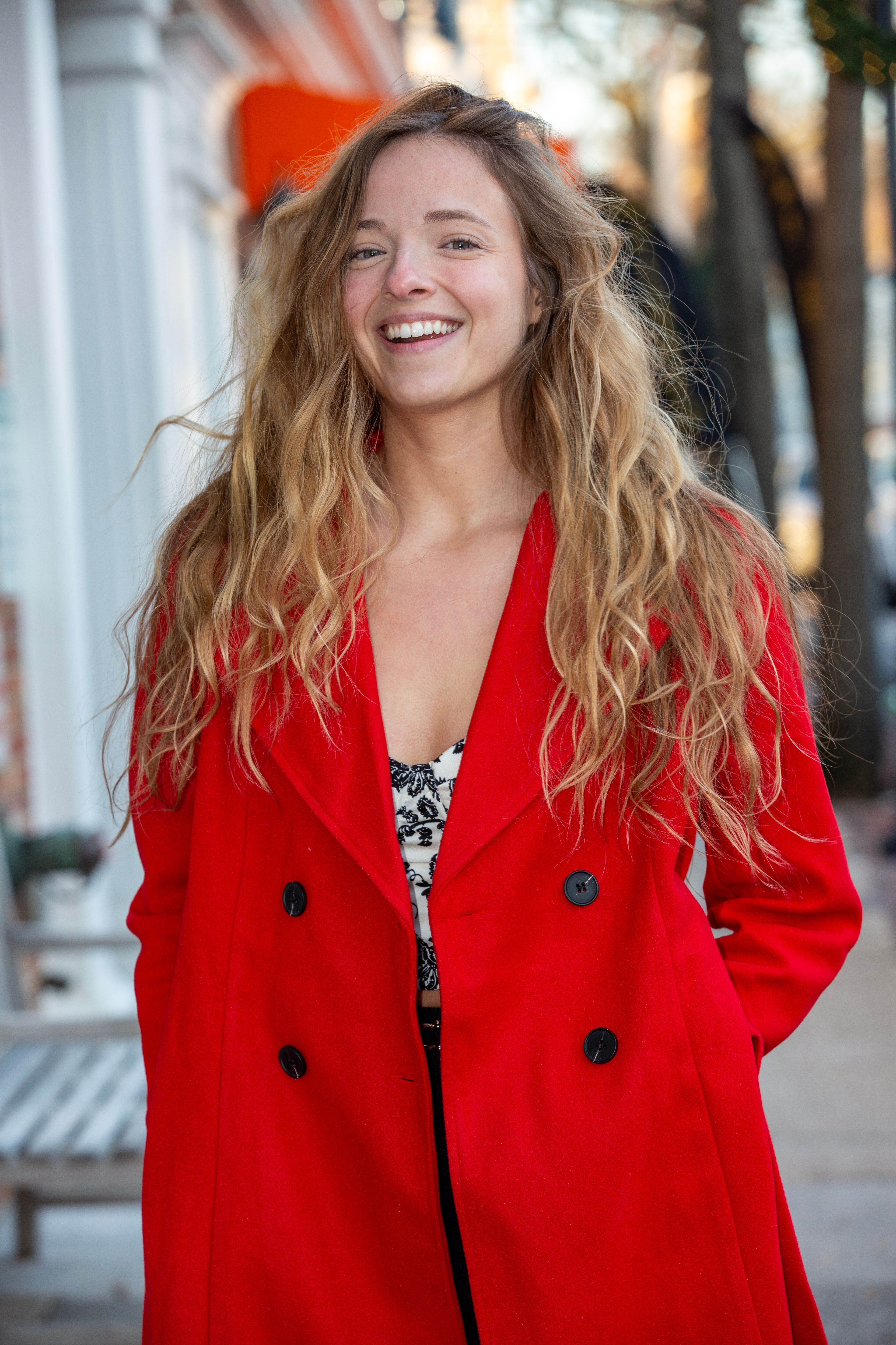 A joyful young woman in a red coat smiling on a sunny street.